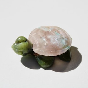 May include: A small, carved stone turtle figurine. The turtle's head and legs are a vibrant green, while the shell is a soft pink with subtle blue-green veining. The shell has carved lines. The figurine is on a white background.