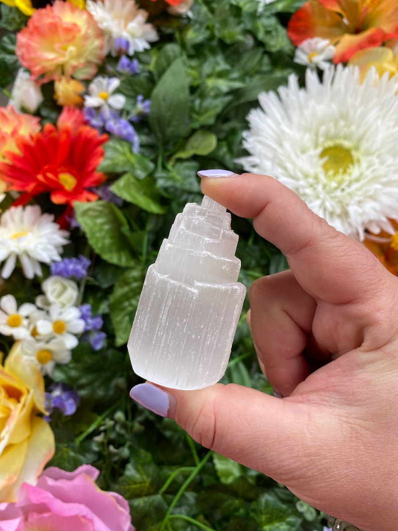 May include: A white selenite crystal tower with a layered, pointed top. The crystal is smooth and has a natural, raw texture.