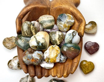 May include: A wooden hand-shaped bowl filled with heart-shaped polished stones in shades of green, brown, and white. The stones are scattered around the bowl on a white surface.