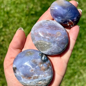 May include: Three polished, oval-shaped stones in shades of blue, purple, and brown are held in a hand. The stones have a smooth, glossy surface with natural patterns and variations. The background is blurred green grass.