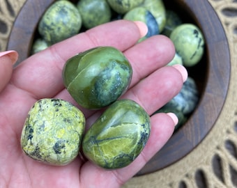 May include: A collection of green and yellow serpentine tumbled stones in a wooden bowl. The stones are smooth and polished, with a variety of patterns and textures.