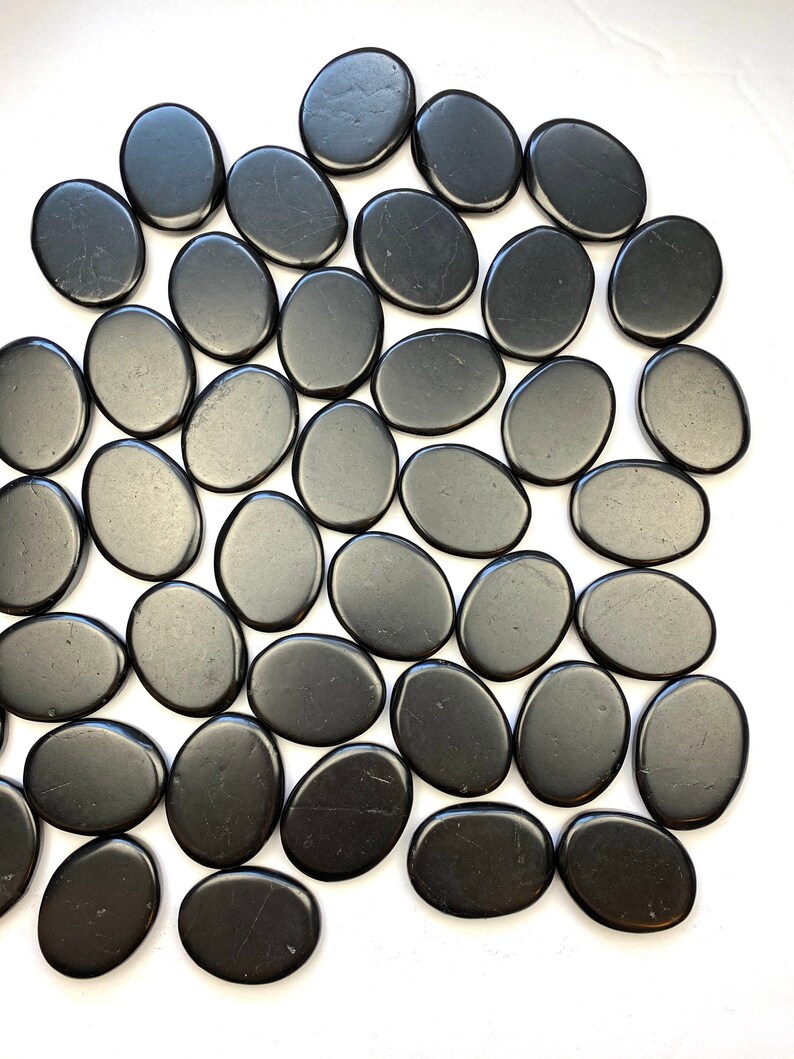 May include: A collection of smooth, black oval-shaped stones. The stones are arranged in a random pattern on a white surface.