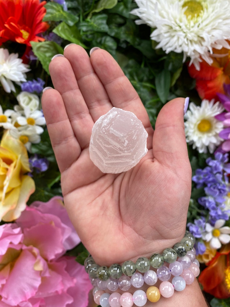 May include: A white selenite crystal with a layered, translucent appearance. The crystal is being held in a hand, surrounded by colorful flowers.