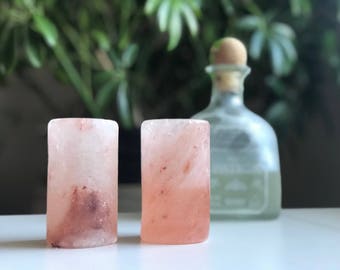 May include: Two pink Himalayan salt shot glasses on a white surface. The glasses are made of natural salt and have a rough texture.