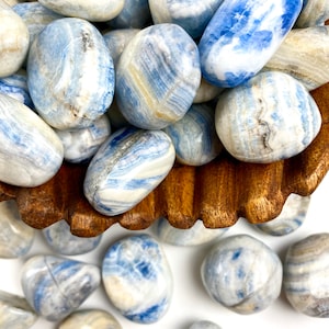 May include: A collection of blue and white banded tumbled stones in a wooden bowl. The stones are smooth and polished, with a variety of blue and white patterns.