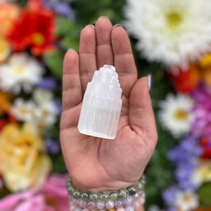 May include: A white selenite tower crystal, also known as a selenite wand, is held in a hand. The crystal is smooth and has a layered, stacked appearance. The background is a blurred image of colorful flowers.