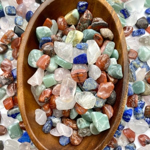 May include: A wooden bowl filled with a variety of polished gemstones in shades of red, blue, green, and clear. The gemstones are tumbled and smooth, with a mix of sizes and shapes. The bowl is oval-shaped and made of a light brown wood.