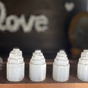 May include: Four white selenite crystal towers of varying sizes, arranged in a row on a wooden surface. The crystals are smooth and have a layered, stacked appearance.
