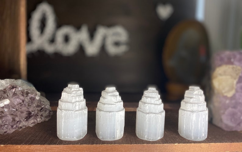 May include: Four white selenite crystal towers of varying sizes, arranged in a row on a wooden surface. The crystals are smooth and have a layered, stacked appearance.