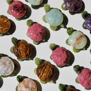 May include: A collection of gemstone turtle figurines in various colors, including pink, brown, purple, and blue. Each turtle has a green base and legs, with a polished, rounded shell. The turtles are arranged on a white surface.