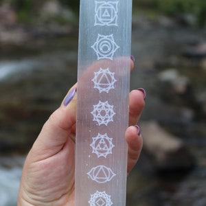 Pu&ograve; includere: Una bacchetta di selenite trasparente con sette simboli di chakra incisi. I simboli sono disposti verticalmente dall'alto verso il basso. La bacchetta &egrave; tenuta in una mano.