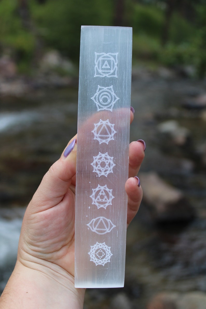 Pu&ograve; includere: Una bacchetta di selenite trasparente con sette simboli di chakra incisi. I simboli sono disposti verticalmente dall'alto verso il basso. La bacchetta &egrave; tenuta in una mano.