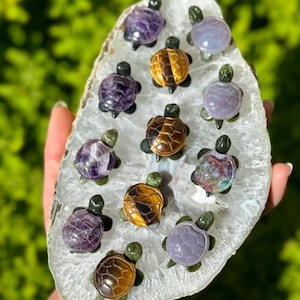 May include: A collection of small, carved gemstone turtle figurines in various colors, including purple, gold, and green. The turtles are displayed on a white and gray geode slice, with a blurred green background.