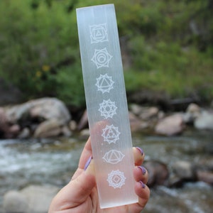 Pu&ograve; includere: Una bacchetta di selenite trasparente con sette simboli dei chakra incisi su di essa. La bacchetta &egrave; tenuta in una mano su uno sfondo sfocato di un ruscello e rocce.