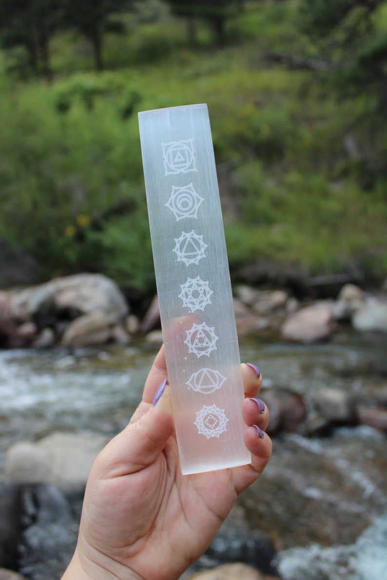 Pu&ograve; includere: Una bacchetta di selenite trasparente con sette simboli dei chakra incisi su di essa. La bacchetta &egrave; tenuta in una mano su uno sfondo sfocato di un ruscello e rocce.
