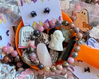 May include: A selection of Halloween-themed pins and charms, featuring ghosts, spiders, and a skull with flowers. The pins are presented in clear plastic bags, alongside pink and brown beads and a small, white ghost figurine. The text "BOO" and "NEVER SLEEP ON A DREAM" can be seen in the image.