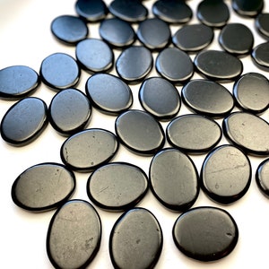 May include: A collection of smooth, black oval-shaped stones. The stones are arranged in a pattern on a white surface.