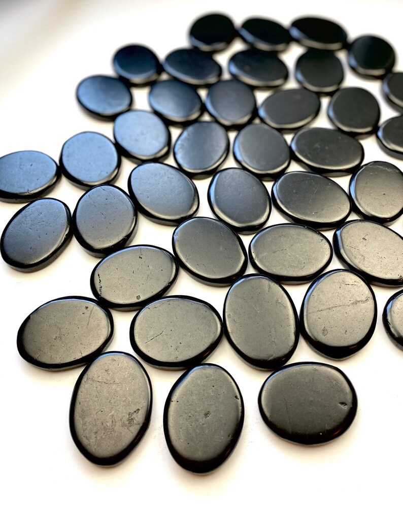 May include: A collection of smooth, black oval-shaped stones. The stones are arranged in a pattern on a white surface.