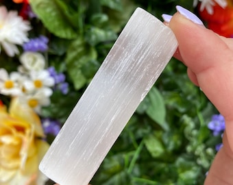 May include: A white selenite crystal wand with a smooth, polished surface and visible striations. The wand is held in a hand against a background of green foliage and colorful flowers.