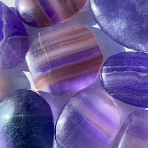 May include: A collection of seven polished fluorite stones in shades of purple and lavender. The stones are smooth and have a natural, striated pattern.