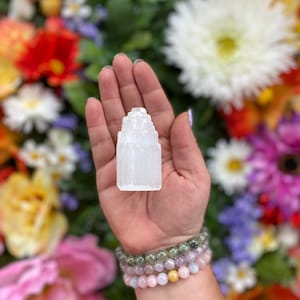 May include: A white selenite crystal tower, also known as a selenite wand, is held in a hand. The crystal is a natural, raw, and unpolished selenite tower. The tower is about 3 inches tall and has a pointed top. The crystal is surrounded by a background of colorful flowers.