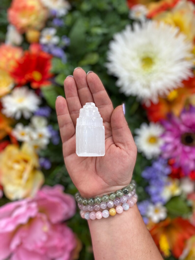 May include: A white selenite crystal tower, also known as a selenite wand, is held in a hand. The crystal is a natural, raw, and unpolished selenite tower. The tower is about 3 inches tall and has a pointed top. The crystal is surrounded by a background of colorful flowers.