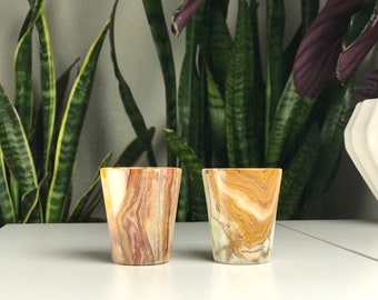 May include: Two small, clear, marble shot glasses with brown and white swirling patterns. The glasses are sitting on a white surface.
