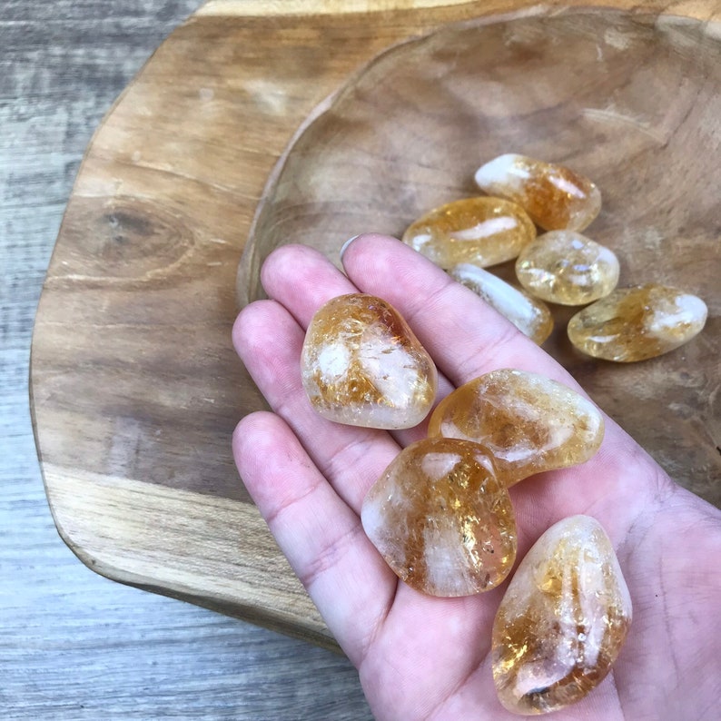 Puede incluir: Una colecci&oacute;n de siete piedras de citrino pulidas, de diferentes tama&ntilde;os y formas, con un color amarillo dorado y una apariencia transl&uacute;cida. Las piedras se muestran en una mano y una superficie de madera.