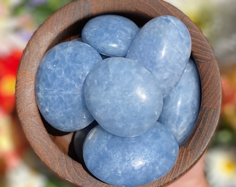 May include: A wooden bowl filled with smooth, light blue tumbled celestite stones. The stones are oval shaped and have a slightly cloudy appearance.