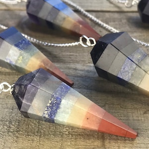 May include: Seven chakra crystal pendulum with a silver chain. The pendulum is made of layered stones in shades of purple, grey, blue, yellow, orange, and red.