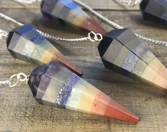 May include: Seven chakra crystal pendulum with a silver chain. The pendulum is made of layered stones in shades of purple, grey, blue, yellow, orange, and red.