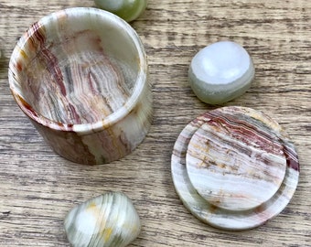 May include: A small, round, white, brown, and red onyx box with a lid. The box is made of natural stone and has a unique, swirling pattern. The box is open and the lid is sitting next to it. There are several small, round, white, brown, and red onyx stones scattered around the box.
