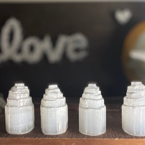 May include: Four white selenite crystal towers, each with a layered, stacked design, arranged in a row on a wooden surface.