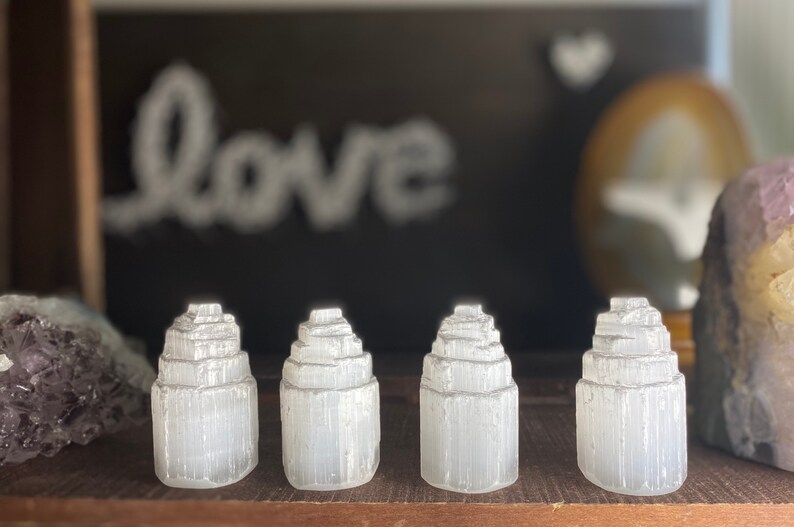 May include: Four white selenite crystal towers, each with a layered, stacked design, arranged in a row on a wooden surface.