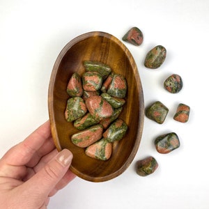 May include: A wooden bowl filled with green and red tumbled stones. The bowl is sitting on a white surface. There are several more stones scattered around the bowl.