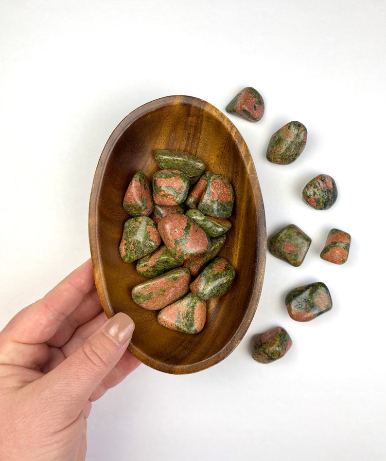 May include: A wooden bowl filled with green and red tumbled stones. The bowl is sitting on a white surface. There are several more stones scattered around the bowl.