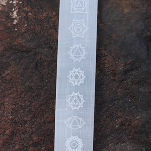 Pu&ograve; includere: Bacchetta di selenite bianca con sette simboli dei chakra incisi sulla superficie. I simboli sono disposti verticalmente dall'alto verso il basso.