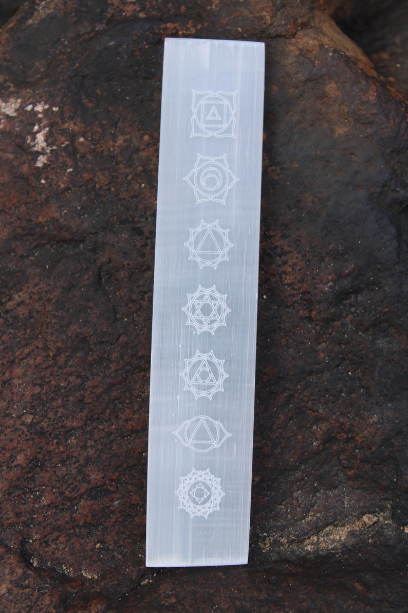 Pu&ograve; includere: Bacchetta di selenite bianca con sette simboli dei chakra incisi sulla superficie. I simboli sono disposti verticalmente dall'alto verso il basso.