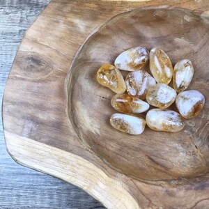 Puede incluir: Un cuenco de madera lleno de una colecci&oacute;n de piedras de citrino lisas, transl&uacute;cidas y de color amarillo dorado.