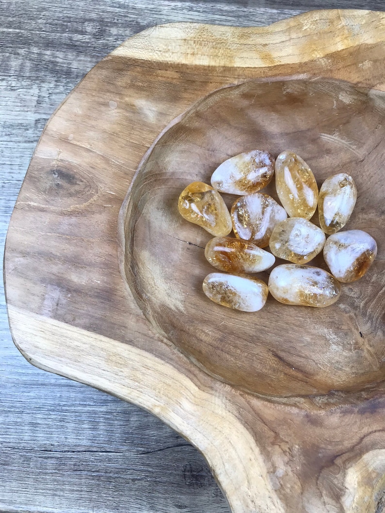 Puede incluir: Un cuenco de madera lleno de una colecci&oacute;n de piedras de citrino lisas, transl&uacute;cidas y de color amarillo dorado.