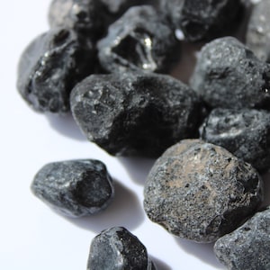 May include: A close-up of a pile of black obsidian stones. The stones are smooth and have a slightly rough texture. They are all different sizes and shapes.