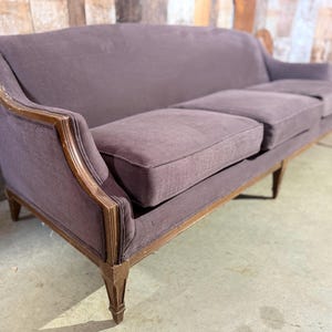May include: A three-seater sofa in a mauve fabric, with a wooden frame. The sofa has decorative detailing on the arms and legs. The cushions are designed for comfort. The sofa's dimensions are suitable for a living room setting.