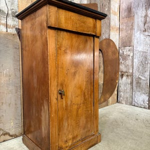 May include: A vintage wooden cabinet with a dark top and a small keyhole. The cabinet is made of light brown wood with visible grain and a rectangular shape. The cabinet has a small door and a decorative trim.