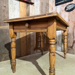 May include: A rectangular wooden table featuring a natural light brown finish with visible wood grain. The table has four turned legs and a single front-facing drawer equipped with a small, round wooden knob centred on the drawer front panel.