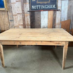 May include: A large, rectangular, light-coloured wooden table with four angled legs. The table has a natural wood grain and appears to be handmade. The table is approximately 183 cm long and 91 cm wide.