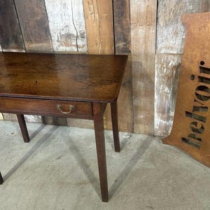 May include: A small, rectangular wooden table with a dark brown finish. It features a single drawer with two brass pulls. The table has four slender legs and is approximately 76 cm long.