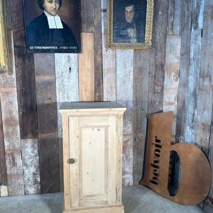 May include: A pale wooden cabinet with a single door and a small brass knob. Two framed portraits are mounted on the wooden plank wall behind the cabinet. A large metal letter "b" with the word "belvoir" cut out is on the right.