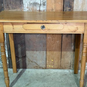 May include: A small, light-coloured wooden table with a single drawer. The table has four legs, each with a turned design. The drawer features a small, round metal knob. The table is approximately 76 cm long.