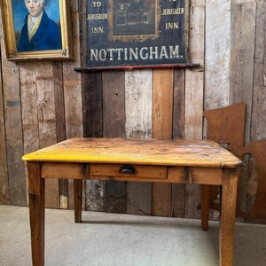 May include: A rectangular wooden table with a natural wood grain finish and a single central drawer featuring a dark metal cup pull handle. The tabletop surface shows signs of wear and a distinct yellow painted edge along the front side.