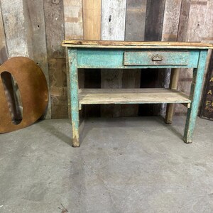 May include: An antique wooden table with a drawer and a shelf, painted in distressed turquoise. The table shows signs of wear. The table measures approximately 76 cm wide, 38 cm deep, and 76 cm high. The word "belvoir" is visible on a metal sign.